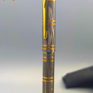 以下が含まれることがあります： 模様のあるグレーとゴールドのボディの高級ペン。クリップとバンドにゴールドのアクセントがあります。「Enigma」の文字がカラフルで様式化されたフォントで表示されています。