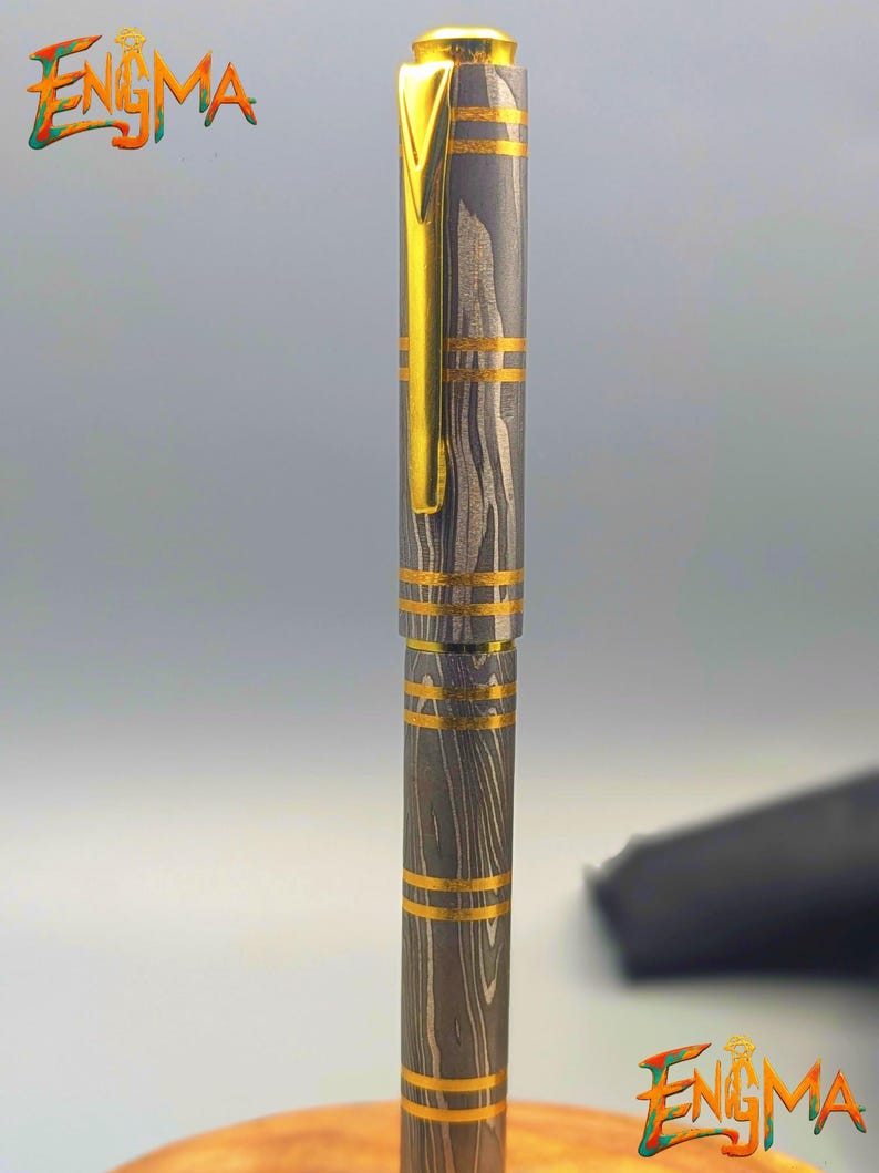 以下が含まれることがあります： 模様のあるグレーとゴールドのボディの高級ペン。クリップとバンドにゴールドのアクセントがあります。「Enigma」の文字がカラフルで様式化されたフォントで表示されています。