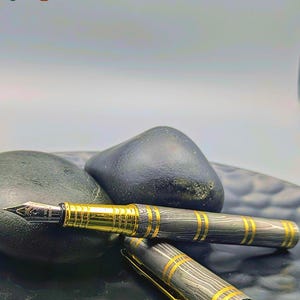 以下が含まれることがあります： 黒と金のデザインの高級万年筆。 ペンは、金色のアクセントと金のペン先を備えた模様のバレルを備えています。 ペンはキャップを横に置いて表示され、左上隅に「Enigma」という単語があります。