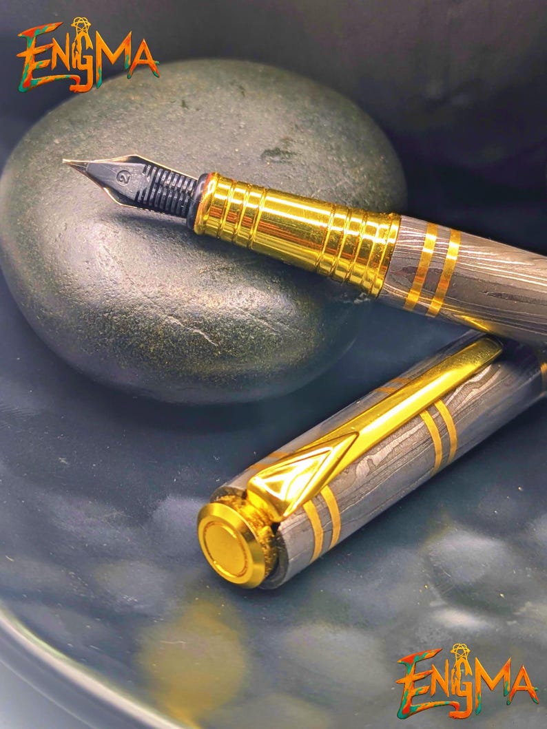 以下が含まれることがあります： 金色のペン先とアクセントが付いた万年筆で、グレーとゴールドのストライプのバレルが特徴です。 ペンはキャップを外して表示され、ペン先が見えます。 「Enigma」という言葉が左上と右下にあります。