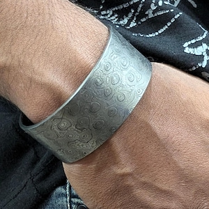 Könnte beinhalten: Ein breites silbernes Armband mit einem strukturierten, wirbelnden Muster. Das Armband ist aus Metall und hat eine polierte Oberfläche.