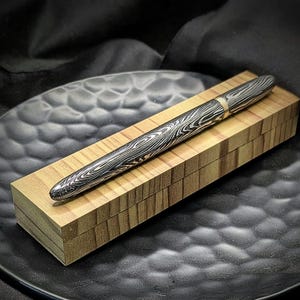 May include: A patterned metal fountain pen and wooden pen box. The pen features a distinctive swirling pattern in silver and black, with a gold-coloured band. It rests on a light brown wooden box, showcasing a natural wood grain pattern.