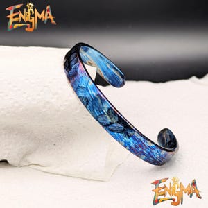Op de afbeelding: Een blauw en paars manchetarmband met een textuurpatroon. De armband heeft het woord "Enigma" in kleurrijke tekst aan de boven- en onderkant van de afbeelding. De armband ligt op een wit oppervlak.