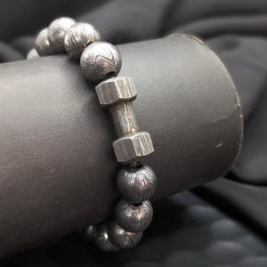 May include: A silver bracelet with a textured metal design. The bracelet is made of round beads and a single, larger, rectangular bead.