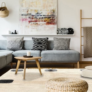 May include: A cream-colored area rug featuring a shaggy texture and evenly spaced dark charcoal gray square patterns. The rug is positioned in a living room setting with a gray sectional sofa, a wooden coffee table, and a woven floor pouf.