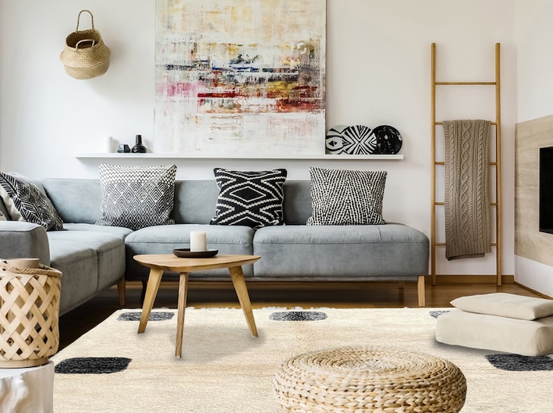 May include: A cream-colored area rug featuring a shaggy texture and evenly spaced dark charcoal gray square patterns. The rug is positioned in a living room setting with a gray sectional sofa, a wooden coffee table, and a woven floor pouf.