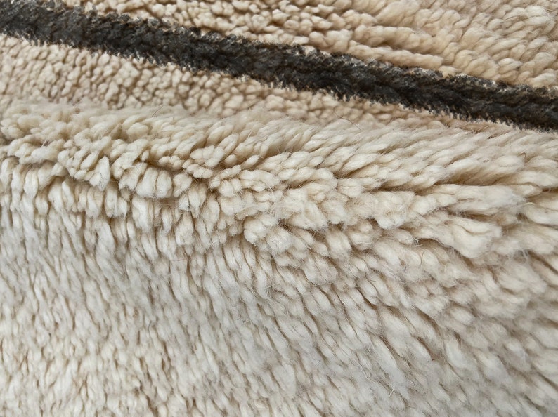 May include: A thick, cream-colored shag rug featuring a single, prominent dark brown horizontal stripe. The texture is dense and plush, with long, soft fibers creating a deep pile surface that shows a slight fold in the fabric.