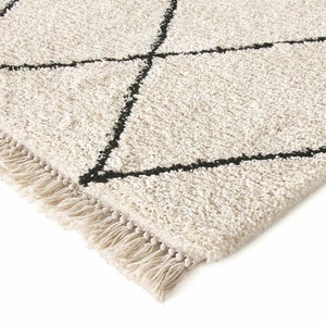 May include: A cream-colored area rug with a high-pile, shaggy texture featuring a black geometric diamond pattern. The rug is finished with a row of short, cream-colored fringe along the edge, providing a tactile and textured appearance.