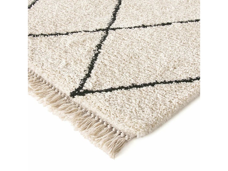 May include: A cream-colored area rug with a high-pile, shaggy texture featuring a black geometric diamond pattern. The rug is finished with a row of short, cream-colored fringe along the edge, providing a tactile and textured appearance.