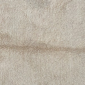 May include: A close-up view of a beige, textured fabric surface featuring a horizontal seam running across the middle. On the far right edge, a portion of a dark, rounded shape is visible, contrasting with the light-colored, fuzzy material.