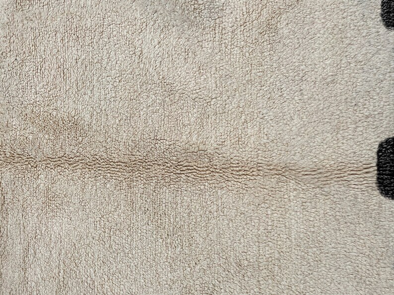 May include: A close-up view of a beige, textured fabric surface featuring a horizontal seam running across the middle. On the far right edge, a portion of a dark, rounded shape is visible, contrasting with the light-colored, fuzzy material.