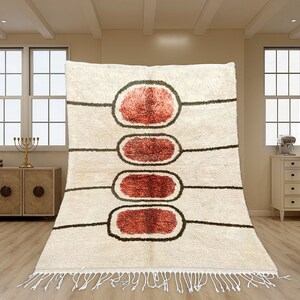 May include: A cream-coloured area rug featuring four vertically aligned, oval-shaped motifs in shades of rust red, each outlined in dark brown. The rug includes horizontal dark brown lines extending from the ovals and white fringe at the bottom.