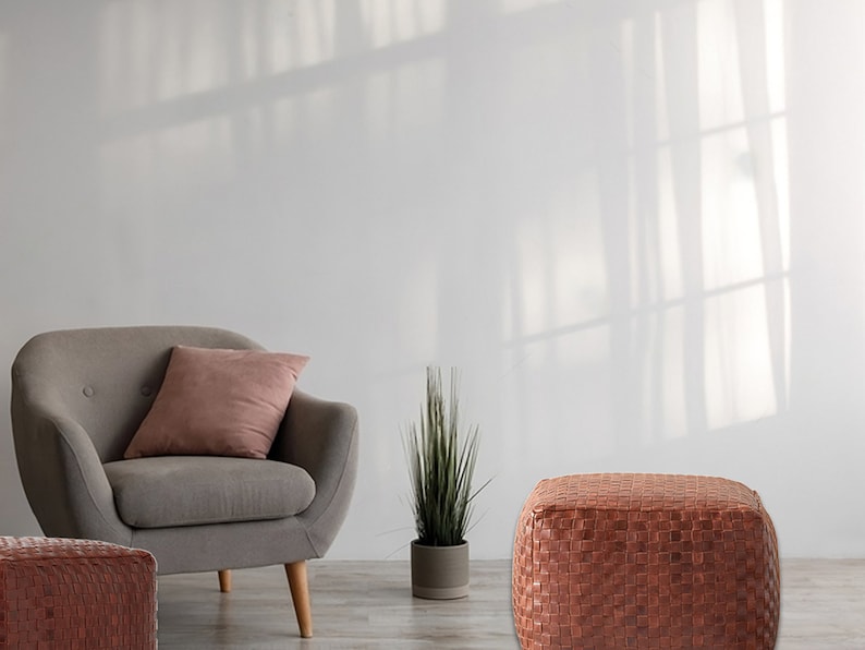 May include: A gray upholstered armchair with light wooden legs and a dusty rose throw pillow, a small potted green plant, and two square, woven brown leather ottomans arranged in a room with light-colored walls.