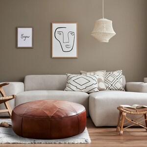 Op de afbeelding: Een ronde, bruine lederen poef met gestikte paneeldetails staat voor een lichtgekleurde hoekbank. De woonkamer bevat een houten stoel, een kleine bamboe kruk met een open boek en twee ingelijste kunstwerken aan de muur.