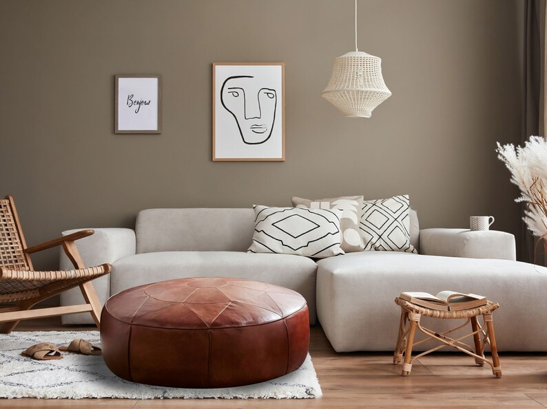 Op de afbeelding: Een ronde, bruine lederen poef met gestikte paneeldetails staat voor een lichtgekleurde hoekbank. De woonkamer bevat een houten stoel, een kleine bamboe kruk met een open boek en twee ingelijste kunstwerken aan de muur.