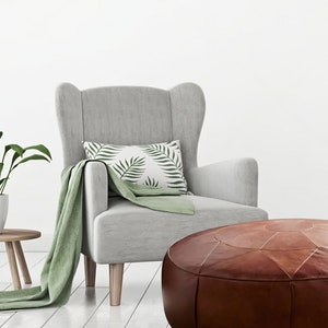 Op de afbeelding: Een lichtgrijze oorfauteuil met lichthouten poten, voorzien van een groene plaid, een wit kussen met groene bladpatronen, een bruine lederen poef en een groene kamerplant in een pot op een kleine houten bijzettafel.