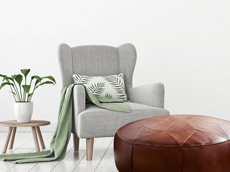 Op de afbeelding: Een lichtgrijze oorfauteuil met lichthouten poten, voorzien van een groene plaid, een wit kussen met groene bladpatronen, een bruine lederen poef en een groene kamerplant in een pot op een kleine houten bijzettafel.
