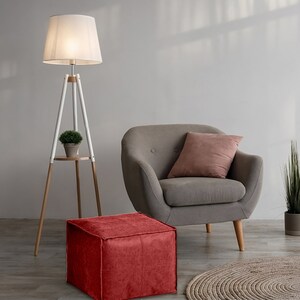 May include: A tripod floor lamp with a white shade and wooden legs, a gray armchair with a dusty pink cushion, a red square ottoman, a small potted plant on the lamp shelf, a tall potted plant, and a round woven rug on the floor.