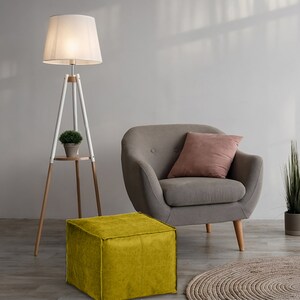 May include: A tripod floor lamp with a white shade and wooden legs, a gray armchair with a pink cushion, a mustard yellow square ottoman, and a small potted plant. The lamp features a circular wooden shelf holding a small green plant.