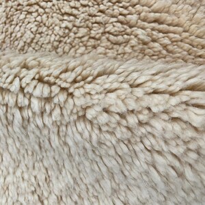 May include: A close-up view of a thick, cream-colored rug with a deep, plush pile. The texture is dense and shaggy, featuring soft, twisted fibers that create a layered, uneven surface with visible folds and natural variations in the weave.