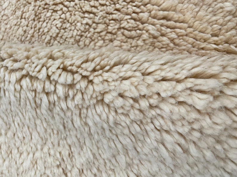 May include: A close-up view of a thick, cream-colored rug with a deep, plush pile. The texture is dense and shaggy, featuring soft, twisted fibers that create a layered, uneven surface with visible folds and natural variations in the weave.