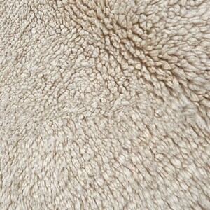 May include: A high-angle close-up view of a thick, cream-colored shag rug with a dense, tufted pile texture. A small portion of a black, textured pattern is visible in the bottom left corner, contrasting with the soft, off-white fibers.