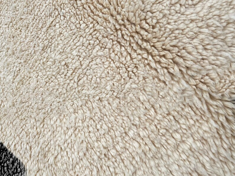 May include: A high-angle close-up view of a thick, cream-colored shag rug with a dense, tufted pile texture. A small portion of a black, textured pattern is visible in the bottom left corner, contrasting with the soft, off-white fibers.