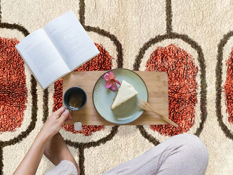 May include: A cream-coloured area rug featuring a repeating pattern of dark brown oval outlines surrounding textured, burnt-orange centres. The rug serves as a backdrop for an open book, a wooden tray, a slice of cake, and a tea mug.