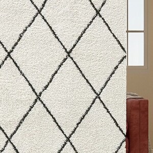 May include: A plush, off-white area rug featuring a repeating black diamond grid pattern. The surface has a thick, textured pile, and the dark lines of the geometric design appear slightly irregular, resembling a hand-drawn or sketched aesthetic.