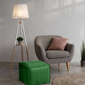 May include: A floor lamp with a white shade and a three-legged wooden base featuring a small circular shelf, a gray armchair with a pink pillow, two green square ottomans, and a potted plant on a circular woven rug.