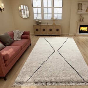 May include: A rectangular, off-white, high-pile area rug featuring a prominent black geometric diamond pattern that spans the center. The rug is finished with short, knotted fringe along the two shorter edges, creating a textured surface.