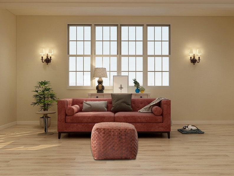 May include: A deep red tufted sofa with bolster pillows and a matching square woven ottoman. The sofa features a dark gray throw pillow and a light gray blanket, positioned in a room with light wood flooring and large windows.