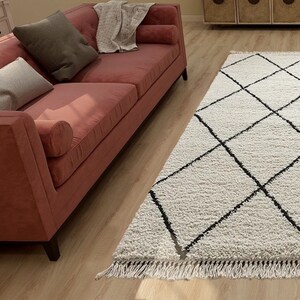May include: A rectangular area rug featuring a white shaggy texture with a black diamond grid pattern and fringed edges. The rug is positioned on a light wood floor next to a coral-colored sofa with two gray throw pillows and a white blanket.