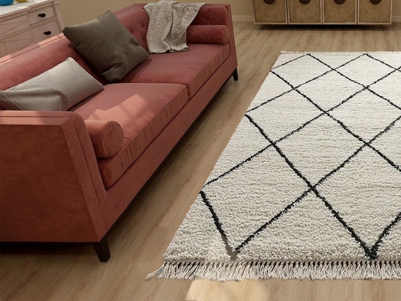 May include: A rectangular area rug featuring a white shaggy texture with a black diamond grid pattern and fringed edges. The rug is positioned on a light wood floor next to a coral-colored sofa with two gray throw pillows and a white blanket.