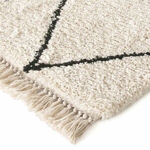 May include: A cream-colored area rug with a thick, shaggy texture and a geometric pattern formed by thin black lines. The rug features a decorative fringe along the edge, consisting of twisted cream-colored yarn tassels.