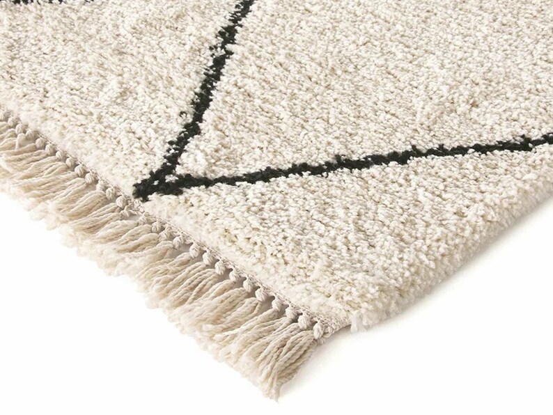 May include: A cream-colored area rug with a thick, shaggy texture and a geometric pattern formed by thin black lines. The rug features a decorative fringe along the edge, consisting of twisted cream-colored yarn tassels.