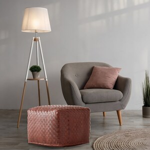 May include: A square, woven, copper-colored leather ottoman with a textured grid pattern. The ottoman is positioned in front of a gray upholstered armchair with a pink pillow, a white tripod floor lamp, and a potted green plant.