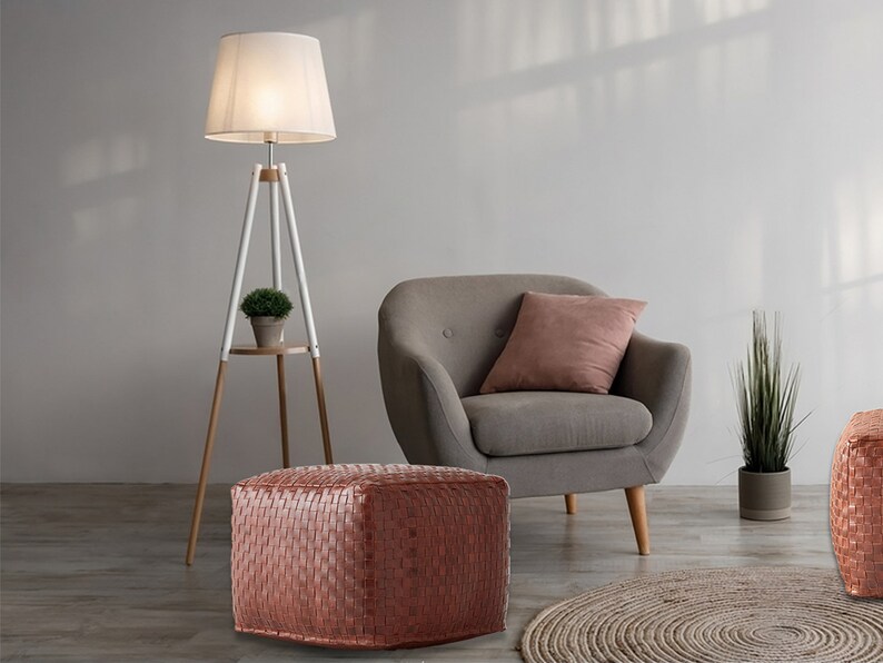 May include: A square, woven, copper-colored leather ottoman with a textured grid pattern. The ottoman is positioned in front of a gray upholstered armchair with a pink pillow, a white tripod floor lamp, and a potted green plant.