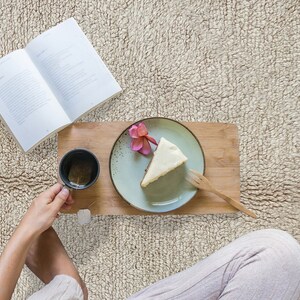 May include: A wooden serving board holds a light green ceramic plate with a slice of cheesecake topped with a pink flower, a wooden fork, and a black mug containing tea with a tea bag, positioned next to an open book.