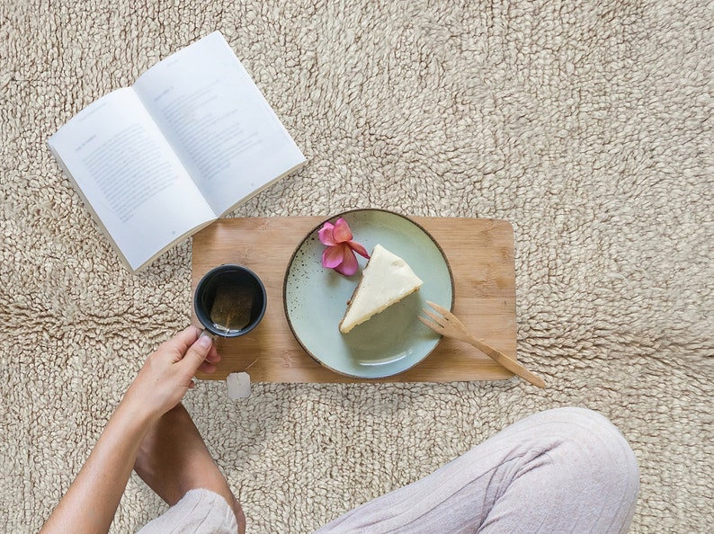May include: A wooden serving board holds a light green ceramic plate with a slice of cheesecake topped with a pink flower, a wooden fork, and a black mug containing tea with a tea bag, positioned next to an open book.