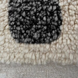 May include: A high-pile textile featuring a thick, cream-colored shag texture surrounding a central, rectangular block of dark charcoal gray, dense, looped wool fibers, set against a flat, woven, light beige fabric base.