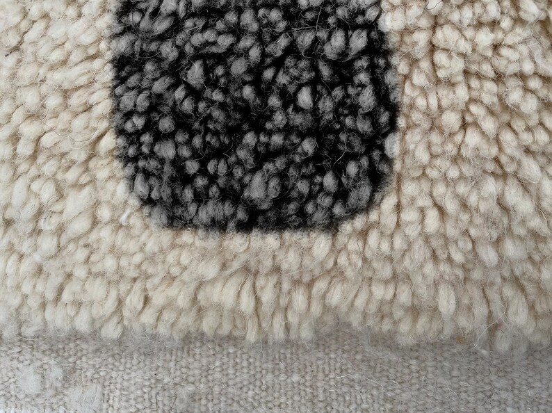 May include: A high-pile textile featuring a thick, cream-colored shag texture surrounding a central, rectangular block of dark charcoal gray, dense, looped wool fibers, set against a flat, woven, light beige fabric base.