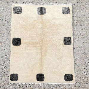 May include: A rectangular area rug featuring a cream-colored base with a high-pile texture. The rug is decorated with eight dark charcoal-colored square motifs arranged in a symmetrical pattern along the edges and includes white fringe.