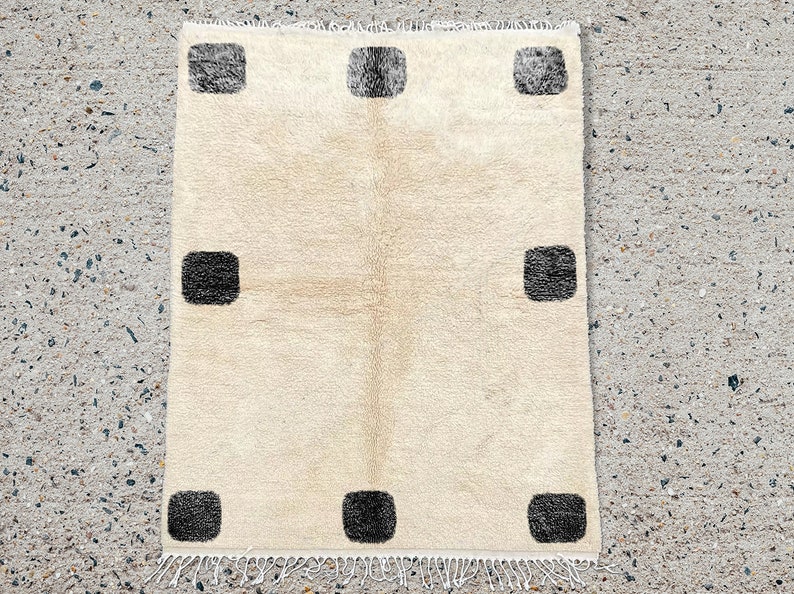 May include: A rectangular area rug featuring a cream-colored base with a high-pile texture. The rug is decorated with eight dark charcoal-colored square motifs arranged in a symmetrical pattern along the edges and includes white fringe.