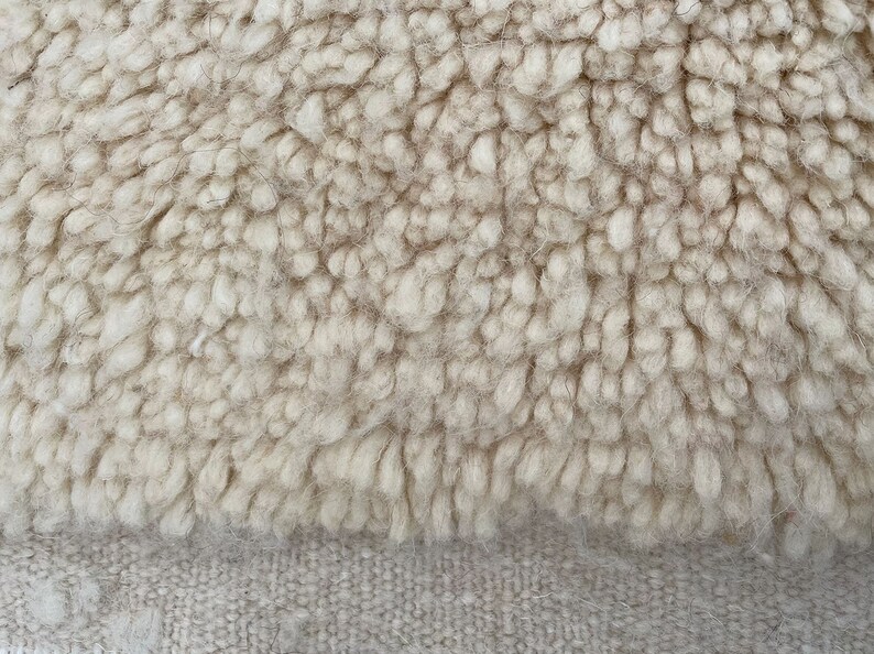 May include: A close-up view of a cream-coloured textile featuring a thick, shaggy pile texture on the upper portion and a flat, tightly woven, light beige fabric base on the lower portion, showcasing the contrast between the two surfaces.