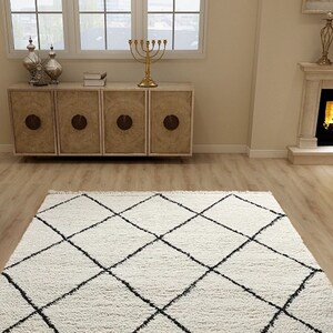 May include: A rectangular area rug featuring a thick, cream-colored shag texture with a prominent black diamond grid pattern. The rug is positioned on a light-toned wood floor, showcasing a soft, plush pile and clean, geometric lines.