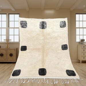 May include: A rectangular area rug featuring a cream-colored base with a repeating pattern of nine dark gray, textured square shapes arranged in a grid. The rug has a thick, plush pile and is finished with white fringe along the bottom edge.