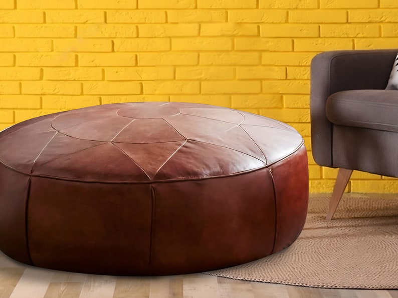 Op de afbeelding: Een ronde, lage bruine lederen poef met een gestikt geometrisch sterpatroon op het bovenoppervlak. Het meubelstuk heeft een gladde, matte afwerking en verticale naden langs de zijkanten, wat zorgt voor een gestructureerde, cilindrische vorm.