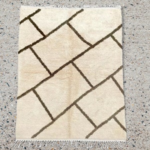 May include: A rectangular area rug featuring a cream-colored base with a dark brown geometric grid pattern of intersecting lines. The rug has a soft, textured pile and includes white fringe detailing along the top and bottom edges.