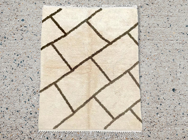 May include: A rectangular area rug featuring a cream-colored base with a dark brown geometric grid pattern of intersecting lines. The rug has a soft, textured pile and includes white fringe detailing along the top and bottom edges.